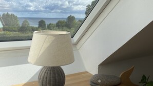 A skylight window with a view of a body of water and trees, a lamp on a wooden table, and a stone sculpture. - House at the sea (Wohlenberg)