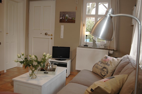 Un salon avec un téléviseur posé sur un meuble blanc, un vase rempli de fleurs sur une table blanche, un canapé beige orné de drapeaux américains et une lampe sur une table d’appoint.
