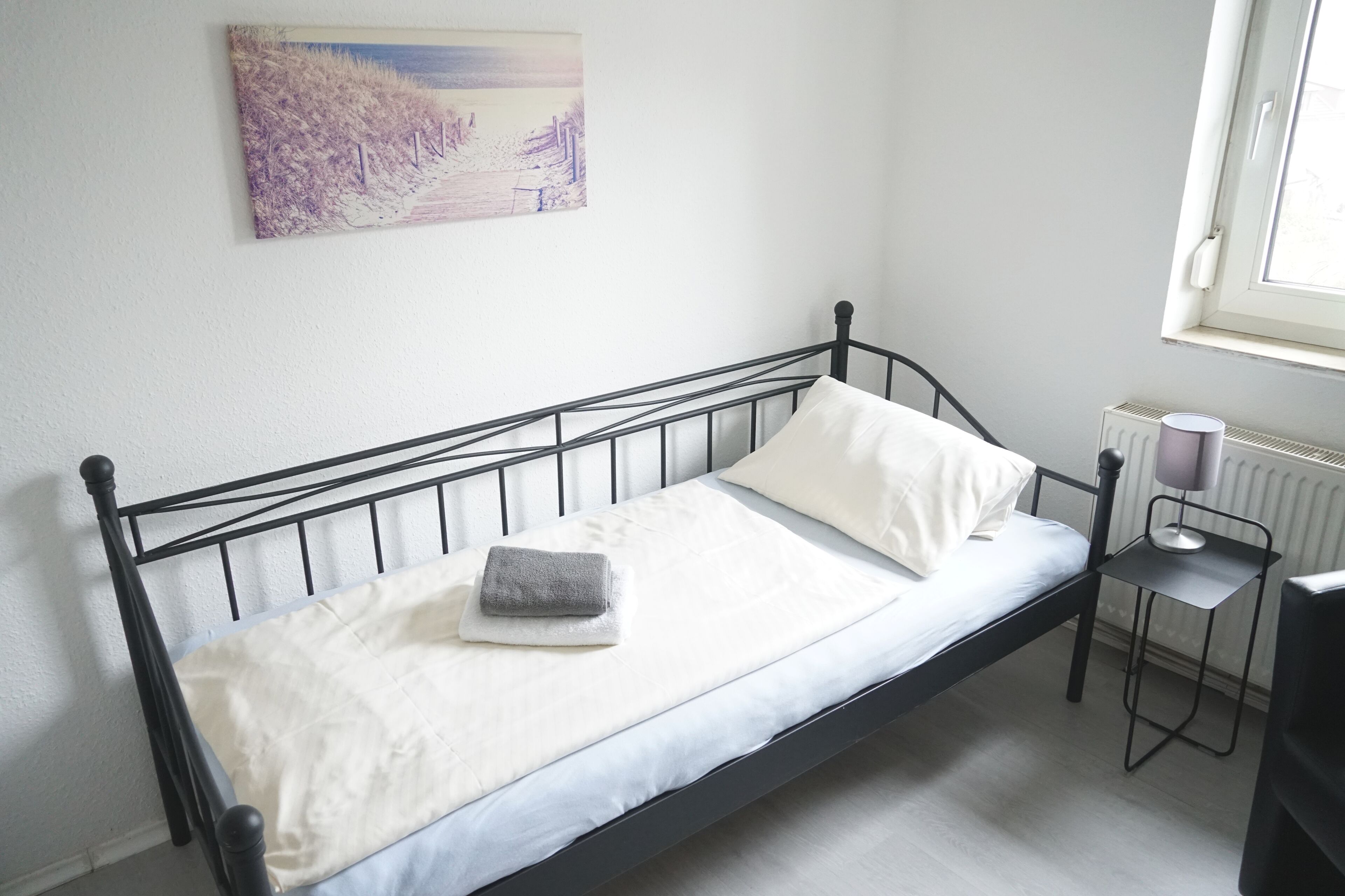 Ein Einzelbett mit weißer Matratze und Kissen, ein kleiner Nachttisch mit Lampe sowie ein Heizkörper.