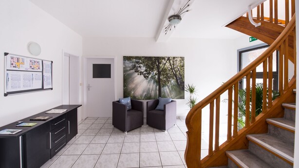 Un intérieur moderne avec un escalier, un coin salon avec deux fauteuils, un meuble noir contenant des papiers et une grande fenêtre donnant sur des arbres.