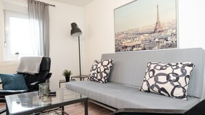 Un salon moderne avec un canapé gris, des coussins à motifs, une table basse en verre, un fauteuil noir, une lampe sur pied et une grande affiche représentant un paysage urbain accrochée au mur.