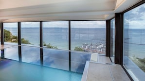 Une piscine intérieure moderne, dotée de grandes baies vitrées offrant une vue sur la mer et sur le paysage urbain au loin.