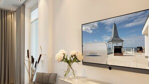 Une chambre d’hôtel moderne dotée d’un grand téléviseur à écran plat fixé au mur, d’un fauteuil confortable, d’un vase avec des fleurs blanches et d’une télécommande posée sur une console blanche.