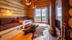 L’intérieur d’une cabane en rondins, avec un plancher en bois, un canapé, une table basse, une cheminée et une grande fenêtre donnant sur un paysage.