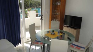 Un coin repas avec une table en verre, des chaises blanches, un téléviseur et une vue sur la mer, visible à travers une porte vitrée.