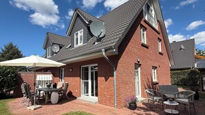 Une maison à deux étages, avec une façade en briques, des antennes paraboliques et une terrasse aménagée avec du mobilier d’extérieur.