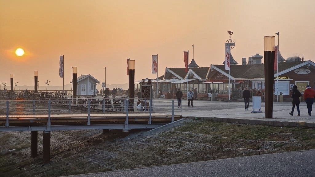 A waterfront promenade with a bridge, buildings, and flags.
