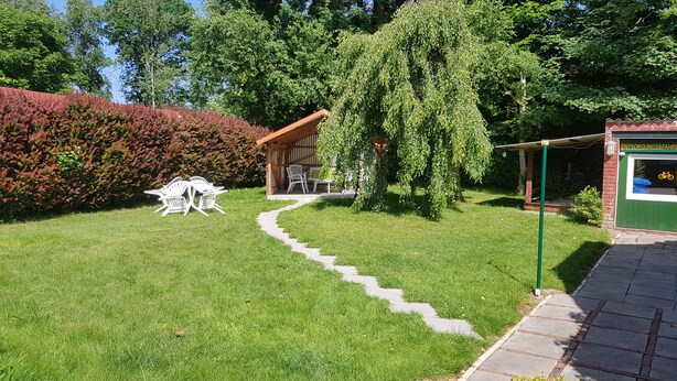 Ein Garten mit einer Holzpergola, einem Weg und einer kleinen Sitzecke mit Stühlen.