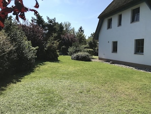 Um edifício branco com telhado de palha, cercado por um gramado bem cuidado e diversos arbustos.