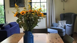 Une pièce avec une table en bois, un vase bleu rempli de fleurs, un canapé bleu, un fauteuil gris et une fenêtre avec des rideaux.
