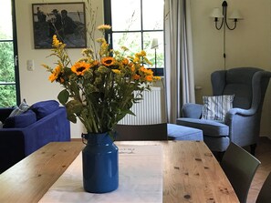 Quarto com mesa de madeira, um vaso azul com flores, sofá azul, poltrona cinza e janela com cortinas.