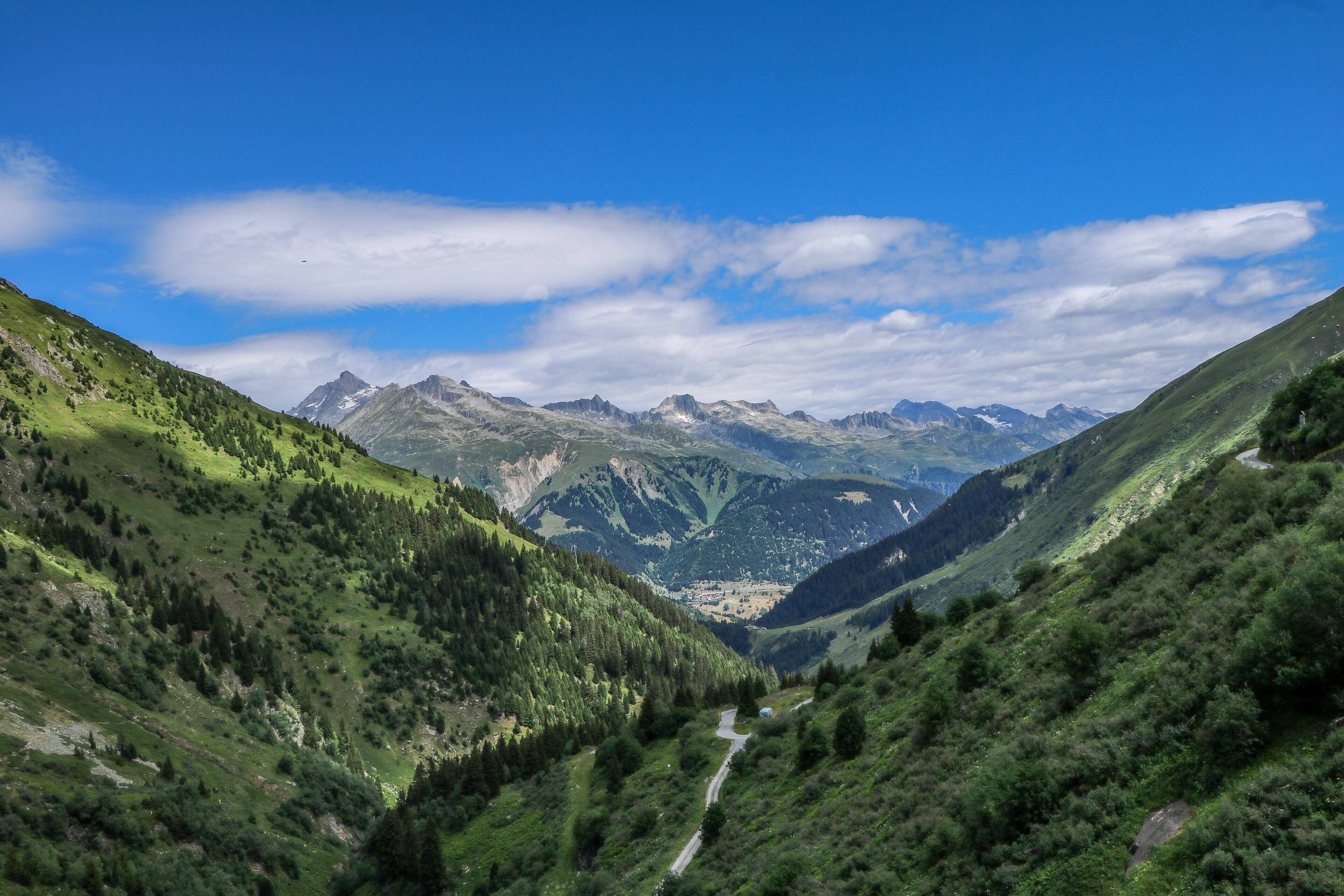 A mountainous landscape with a winding road, lush greenery, and distant peaks under a clear sky.