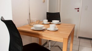 A small dining area with a wooden table, four white cups, bowls, and a glass bowl. There is a black chair with a mesh backrest and a black cushioned seat. The room has white walls and a door with a red handle. - Your little break at the North Sea: modern furnished apartment with living area (Carolinensiel)