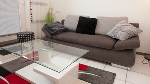A modern living room with a grey sofa, two black and one grey throw pillow, a glass coffee table, and a patterned ottoman. - Your little break at the North Sea: modern furnished apartment with living area (Carolinensiel)
