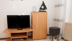 A living room with a flat-screen TV on a wooden stand, a small cabinet, a patterned stool, and a radiator. - Your little break at the North Sea: modern furnished apartment with living area (Carolinensiel)