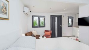 Quarto de hotel moderno com cama, sofá, cadeira, mesinha e TV. Há janelas e um ar condicionado.