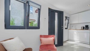 Uma sala de estar moderna com um sofá, uma poltrona vermelha, uma mesa de centro e uma área de cozinha com armários brancos e bancada preta.