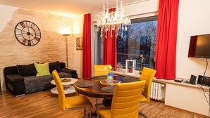 Een moderne woonkamer met een houten vloer, een ronde eettafel met gele stoelen, een zwarte bank met groene kussens, een wandklok en een groot raam met rode gordijnen.