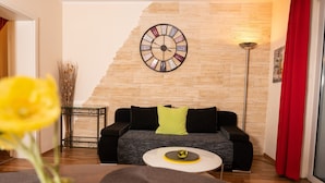 Een moderne woonkamer met een houten wand, een ronde klok, een zwarte bank met groene kussens, een bijzettafeltje met een geel object, en een vaas met gele bloemen.