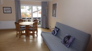 A room with a wooden dining table and chairs, a grey sofa with two cushions, a window with curtains, and a radiator. - Apartment with a beautiful mountain view (Garmisch-Partenkirchen)