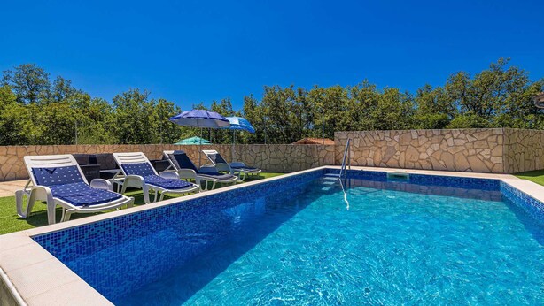 Ein Swimmingpool mit blauen Fliesen, umgeben von Liegestühlen und Sonnenschirmen, vor einer Steinmauer und Bäumen.