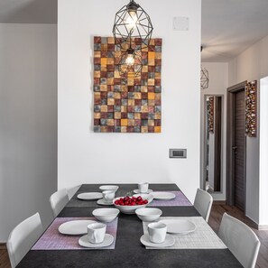 Un coin repas avec une table rectangulaire dressée pour quatre personnes, des chaises blanches, un bol de cerises rouges et un luminaire au design géométrique suspendu au-dessus.