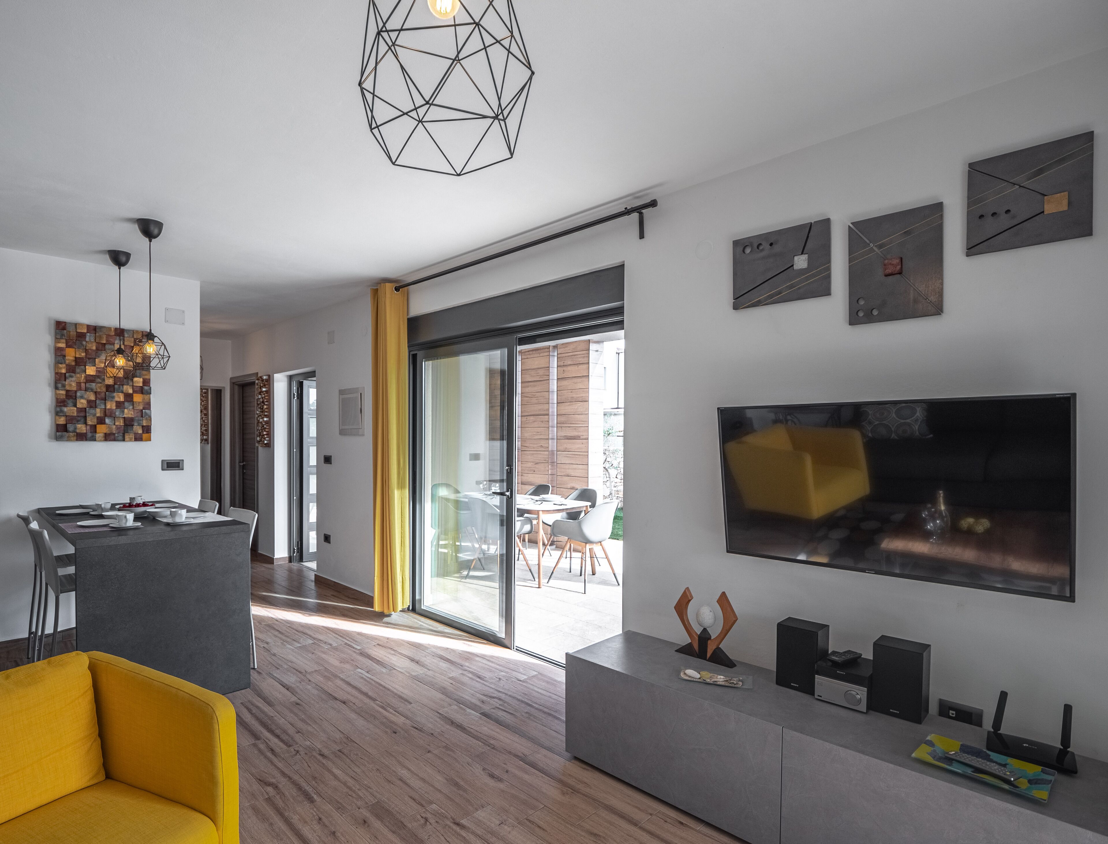 A modern living room with a large flat-screen TV, a yellow sofa, and a dining area with a geometric light fixture.