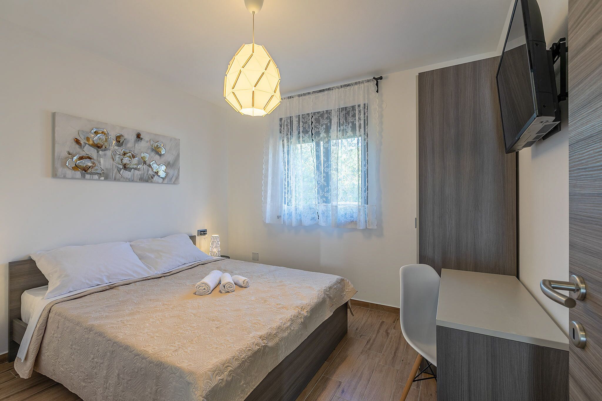 A bedroom with a bed, a desk, a chair, a TV, and a window with curtains.