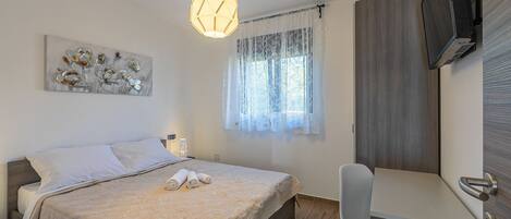 A bedroom with a bed, a desk, a chair, a TV, and a window with curtains.