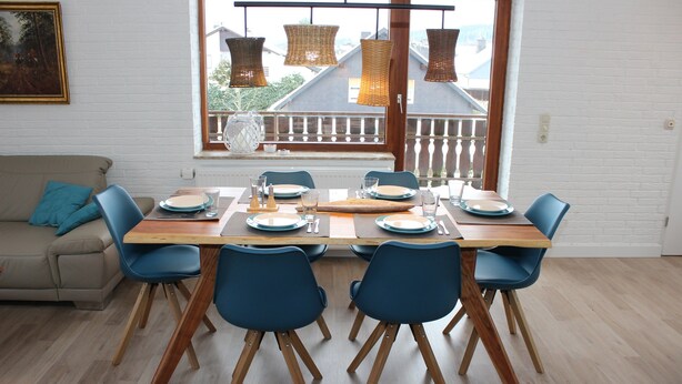 A dining area with a wooden table set for four, blue chairs, and a view of a balcony through a large window. - modern, very spacious apartment in the immediate vicinity of the Center Parcs (Medebach)