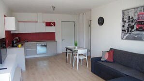 A modern kitchen with white cabinets and a red accent wall, a dining area with a table and chairs, and a living room with a sofa and a wall clock. - Nice vacation apartment in S. Alessandro-Riva del Garda. (riva del garda)