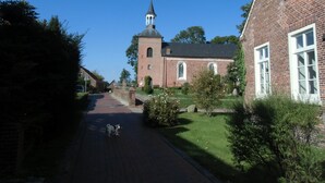 Een gebouw van baksteen met een toren en boogvensters, omringd door groen en een verhard pad.