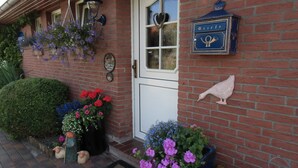 Een gebouw van baksteen met een witte deur, een blauw bord met een vogel en hoorns, en verschillende bloempotten en decoraties.