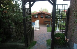A wooden gate leading to a brick pathway, a wooden building with a red roof, and a white chair outside. - Apartment Heimathafen in Werdum near Neuharlingersiel (Werdum)
