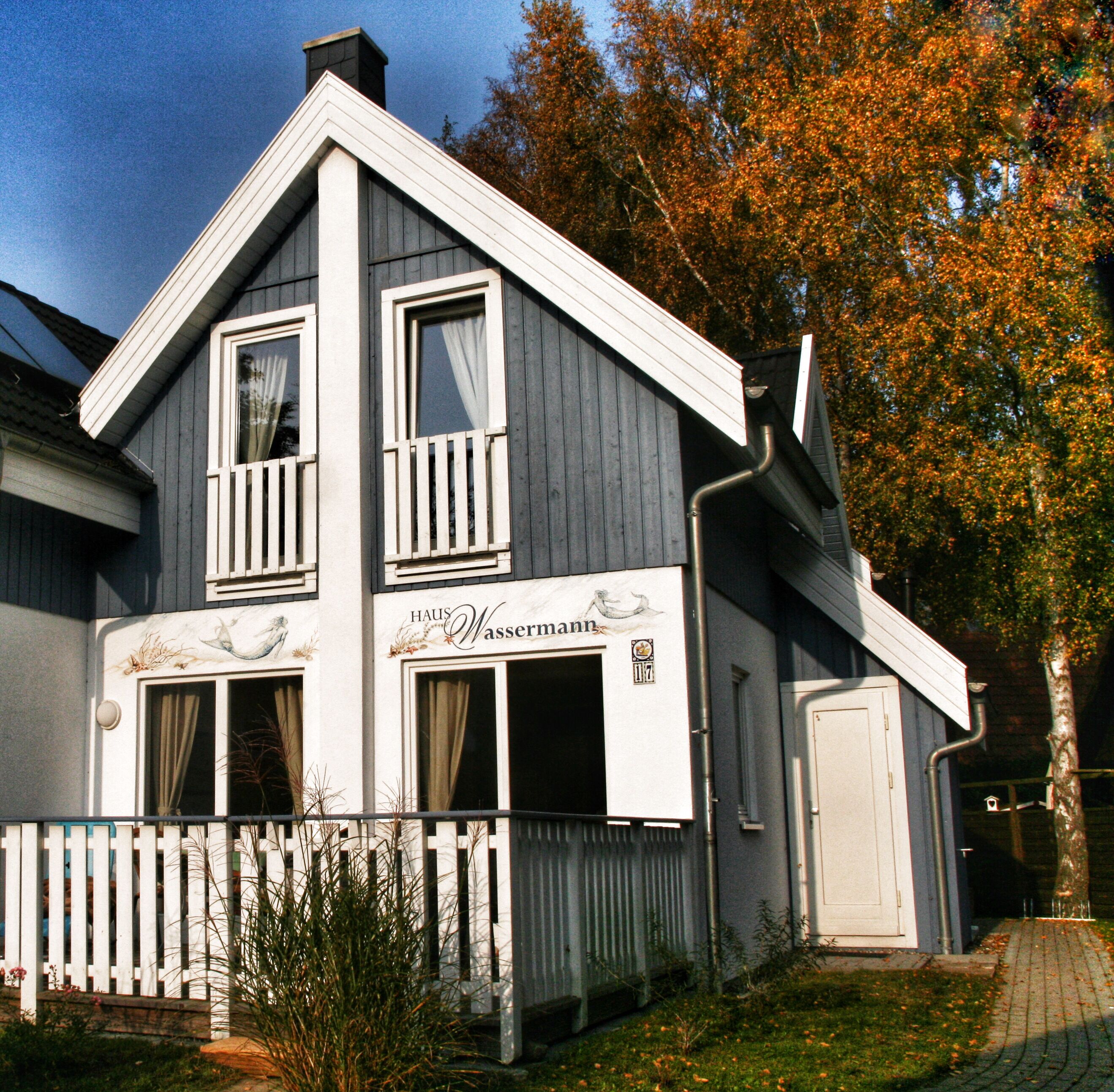 A two-story house with a balcony, a sign reading "Haus Wassermann," and a white picket fence.