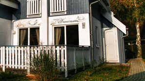 Ein zweistöckiges Haus mit Balkon, einem Schild mit der Aufschrift 'Haus Wassermann' und einem weißen Zaun.