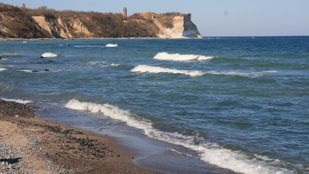 Eine Küstenlandschaft mit Sandstrand, Wellen und einer Klippe mit Leuchtturm.