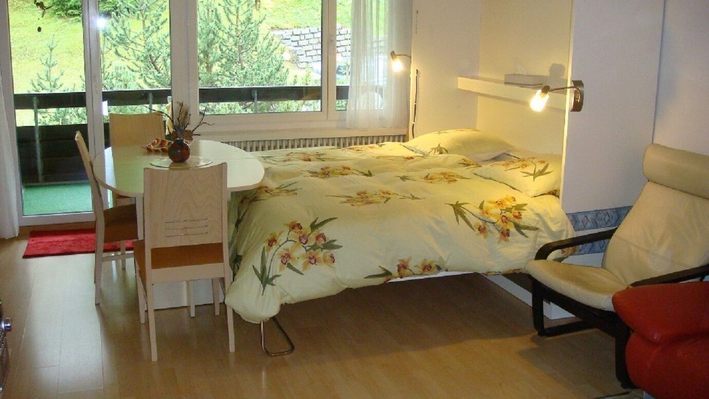 Ein Schlafzimmer mit Bett, Schreibtisch und Stuhl. Ein Fenster bietet Blick auf Grünflächen.