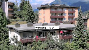 A multi-story residential building with balconies, surrounded by trees and mountains.