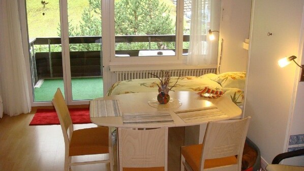 A room with a bed, a table with chairs, a window with a view of greenery, and a radiator.