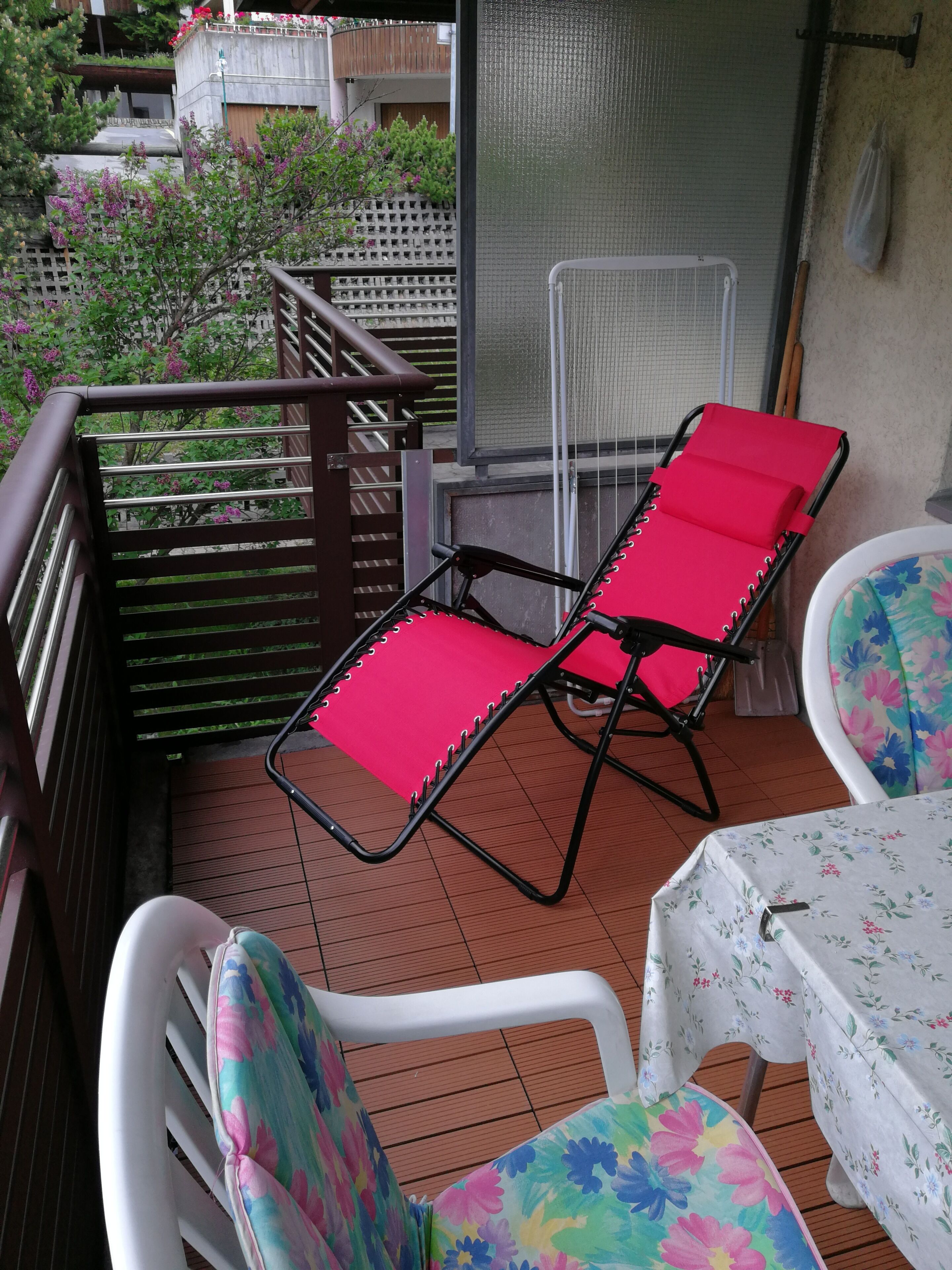Ein Balkon mit zwei roten Liegestühlen, einem weißen Tisch mit einer blumigen Tischdecke und einem weißen Stuhl mit einem farbenfrohen floralen Sitzkissen.