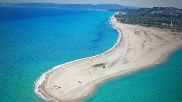 Eine Vogelperspektive zeigt einen Strand mit klarem türkisblauem Wasser, einem sandigen Ufer und einigen Menschen, die die Sonne genießen.