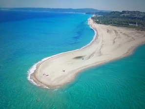 Une vue aérienne d’une plage aux eaux turquoise et limpides, avec un rivage sablonneux et quelques personnes qui profitent du soleil.