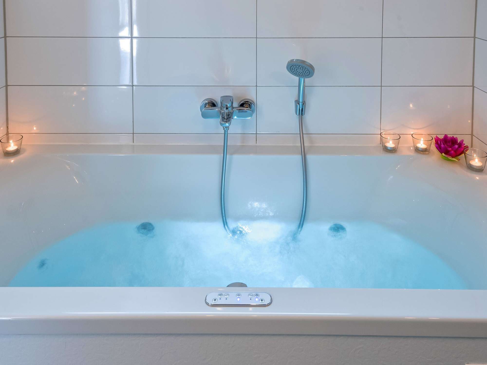 Una bañera moderna con un cabezal de ducha manual y un grifo fijo, rodeada de azulejos blancos y velas.
