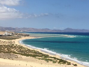 Paisagem costeira com praia arenosa, mar azul cristalino e montanhas ao longe.