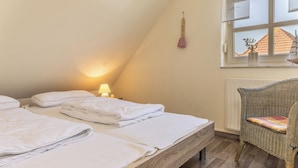 Un dormitorio con dos camas, una mesita de noche con lámpara, una silla de mimbre y una ventana con vistas a un edificio.