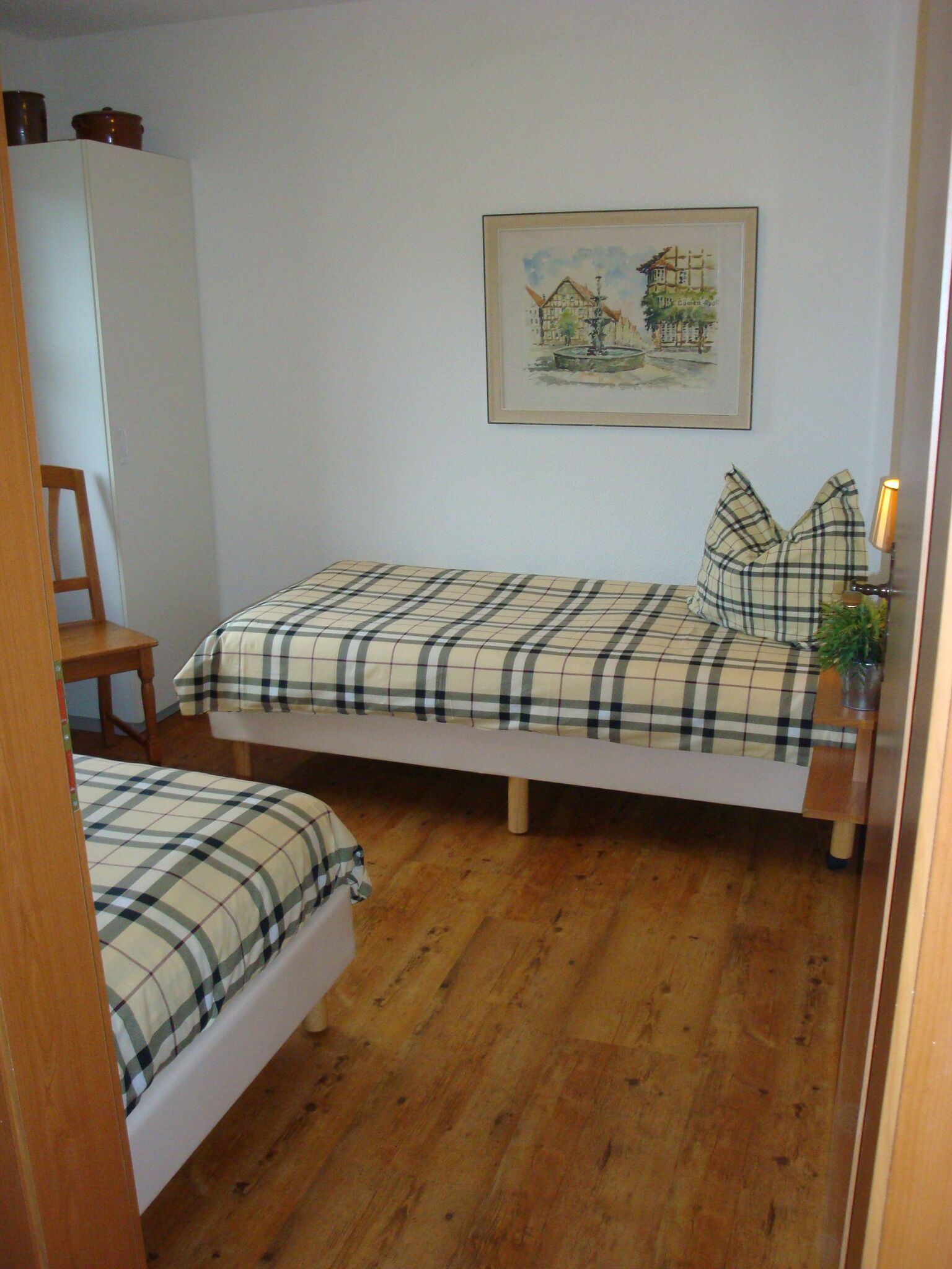Una habitación pequeña con dos camas, piso de madera, un armario blanco y un cuadro enmarcado en la pared.