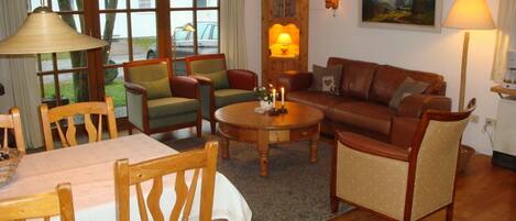 Een gezellige woonkamer met een houten vloer, een bruine leren bank, groene en rode fauteuils, een houten salontafel en een eettafel met stoelen.