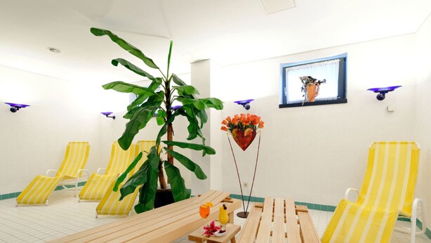 A sunlit room with yellow-striped chairs, a wooden bench, a potted plant, and a wall-mounted TV. - Double room - Hotel Carat (Erfurt)