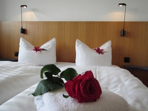 Une chambre d’hôtel avec un lit parfaitement fait, deux oreillers ornés de motifs floraux, une rose rouge et deux lampes fixées au mur.